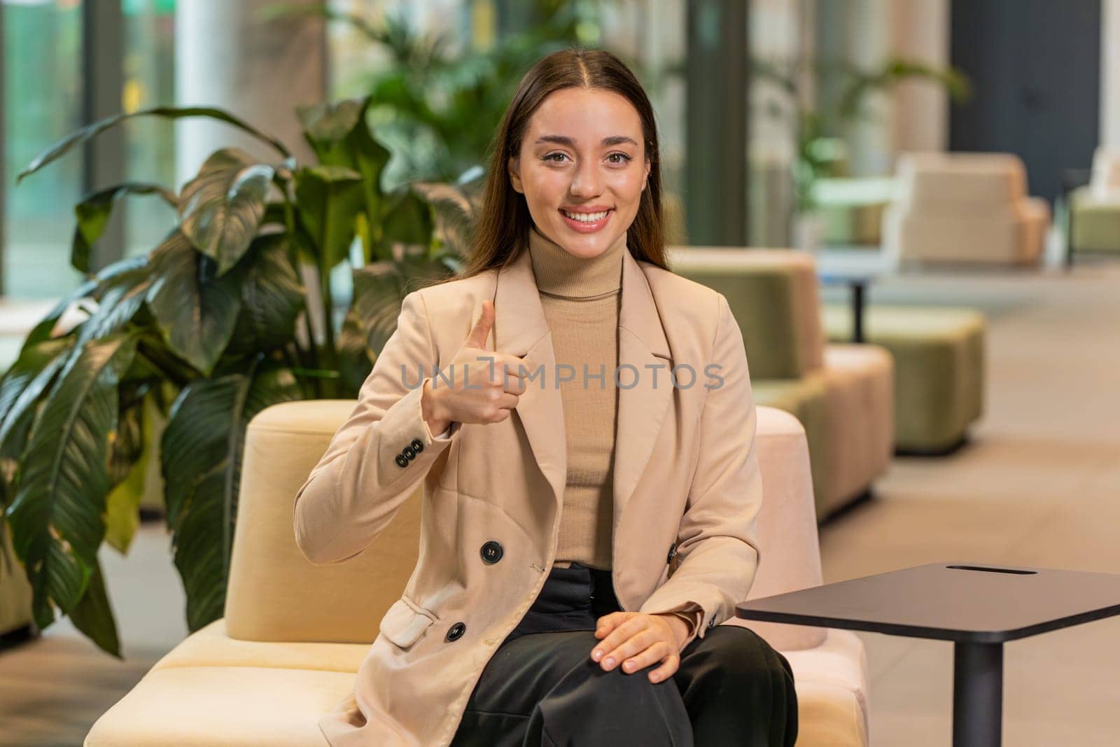 Like. Happy overjoyed young Caucasian woman excited winner celebrate victory success, looking at camera showing thumbs up sitting in office lobby. Portrait of girl employee in formals in hotel hall.