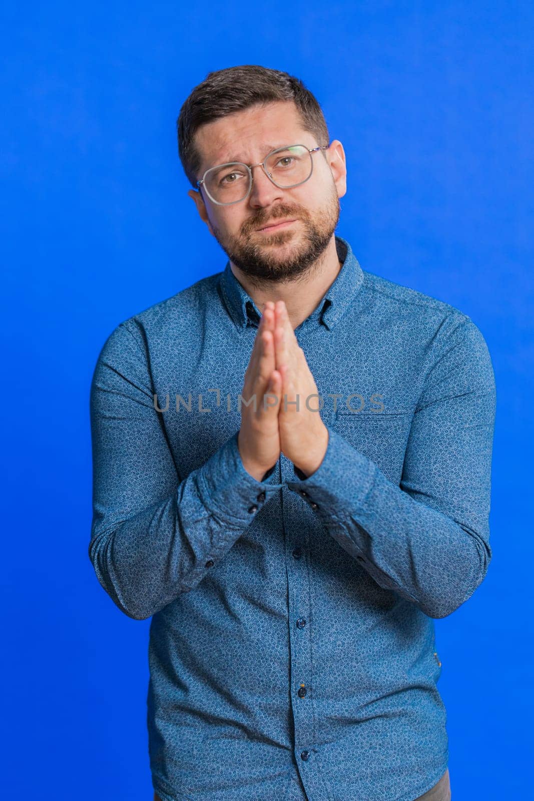Please, God help. Caucasian middle-aged man praying, looking at camera and making wish, asking with hopeful imploring expression, begging apology, meditation. Adult guy isolated on blue background