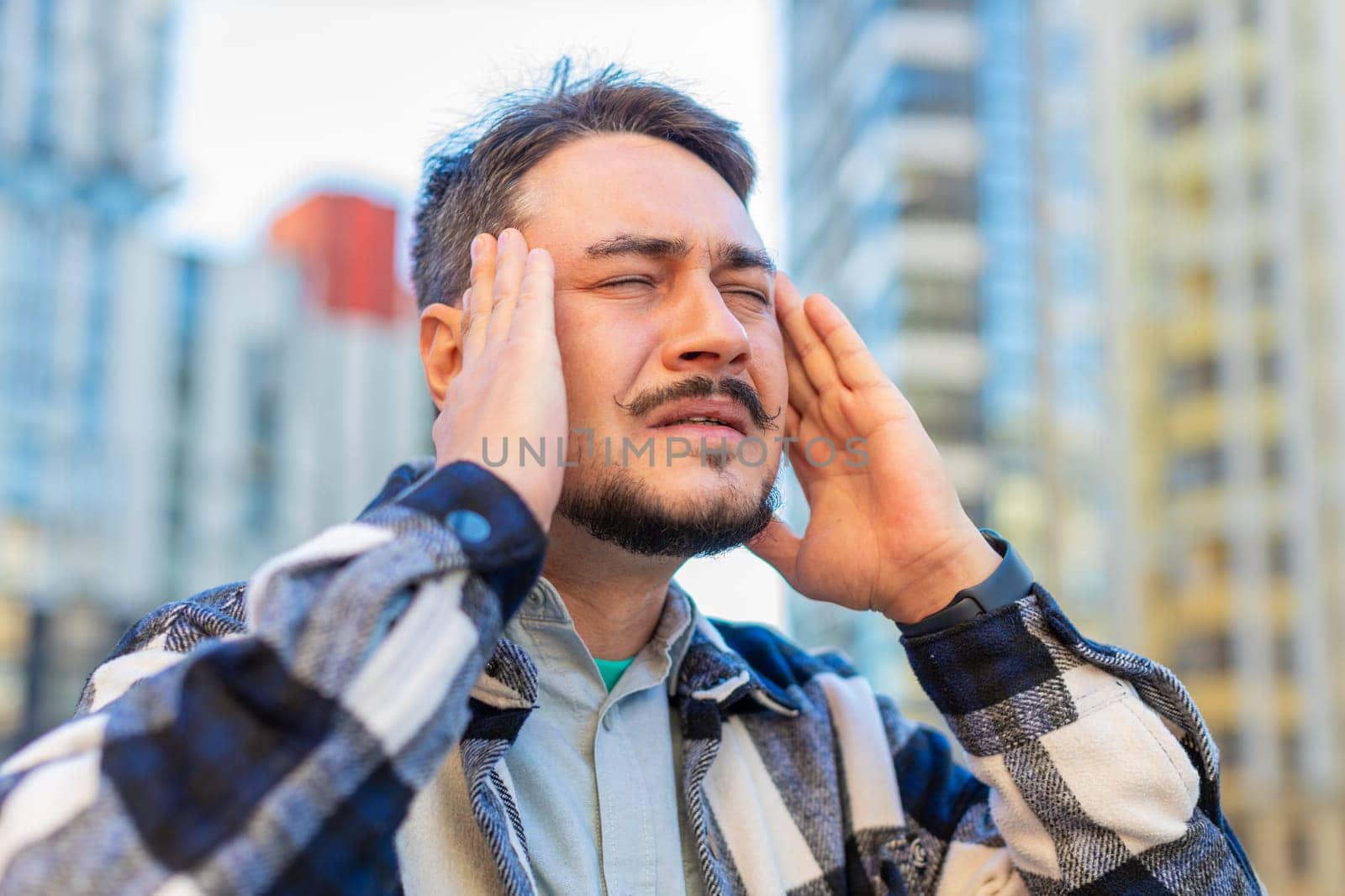 Sick mature Caucasian man feels headache on downtown city street. Middle-aged tourist guy massages her forehead and temples with his hands against building. Upset businessman suffering from migraine.