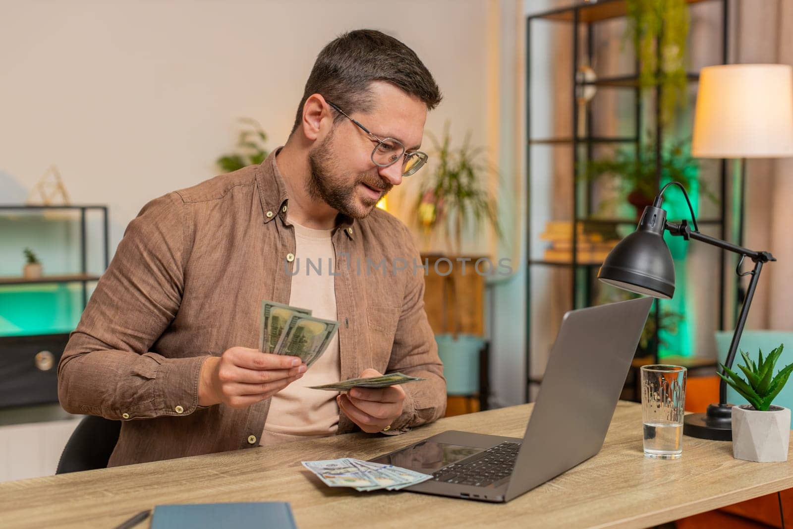 Planning budget. Rich happy businessman counting money dollar cash using laptop calculate domestic bills at home table. Caucasian guy satisfied of income salary earnings for planned vacation gifts.