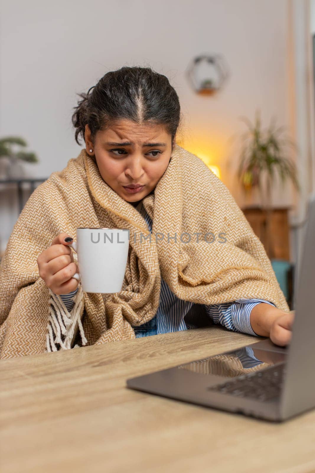 Sick ill Indian young woman freelancer working on laptop wrapped in plaid sit shivering from cold drinking hot coffee in unheated home office. Unhealthy girl trying to warming up in room. Vertical.