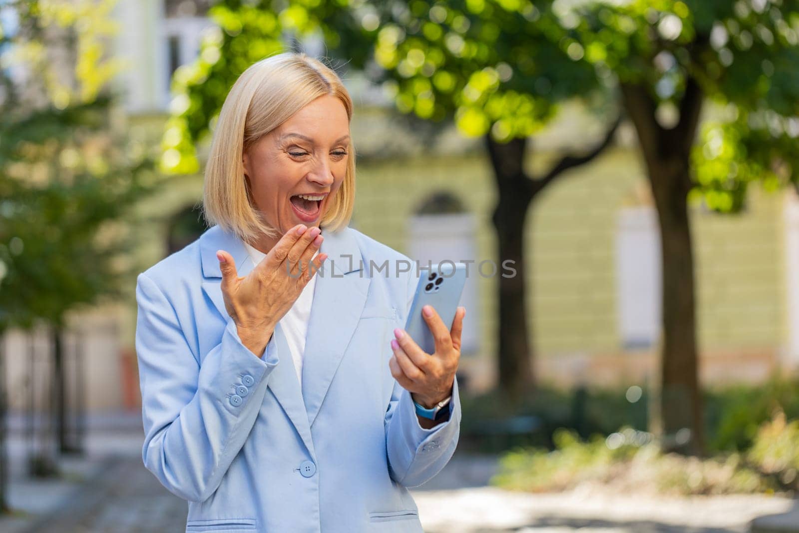 Happy shocked excited Caucasian mature businesswoman in formal suit mouth opened celebrate victory, achievement holding mobile phone good news win message rejoice business success outside city street.