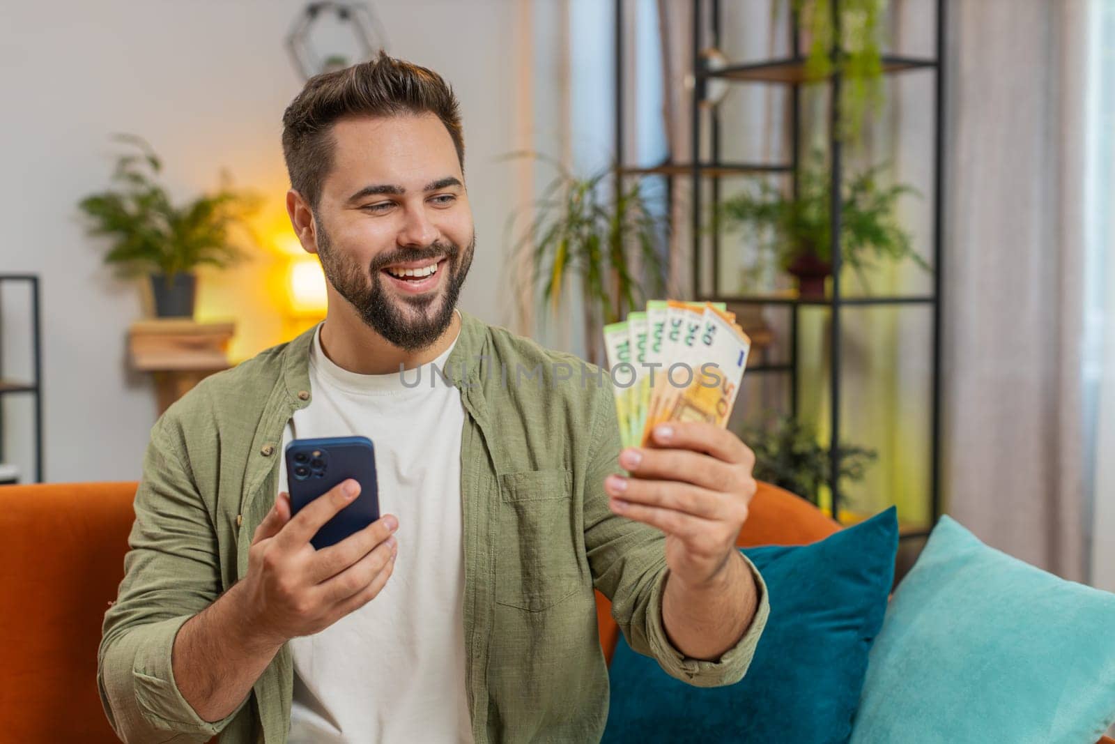 Successful rich young Caucasian man holding and waving euro bill money fan sitting on sofa couch at home. Happy adult guy winning online lottery game planning vacation calculating budget concept.