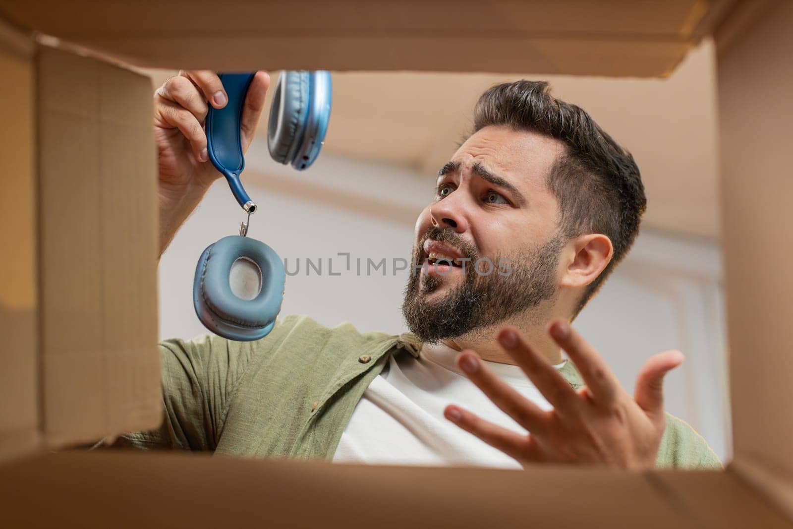 Dissatisfied annoyed male customer open cardboard box receive damaged broken wireless headphones, shocked young man consumer having problem complaint frustrated with bad order delivery at home room.