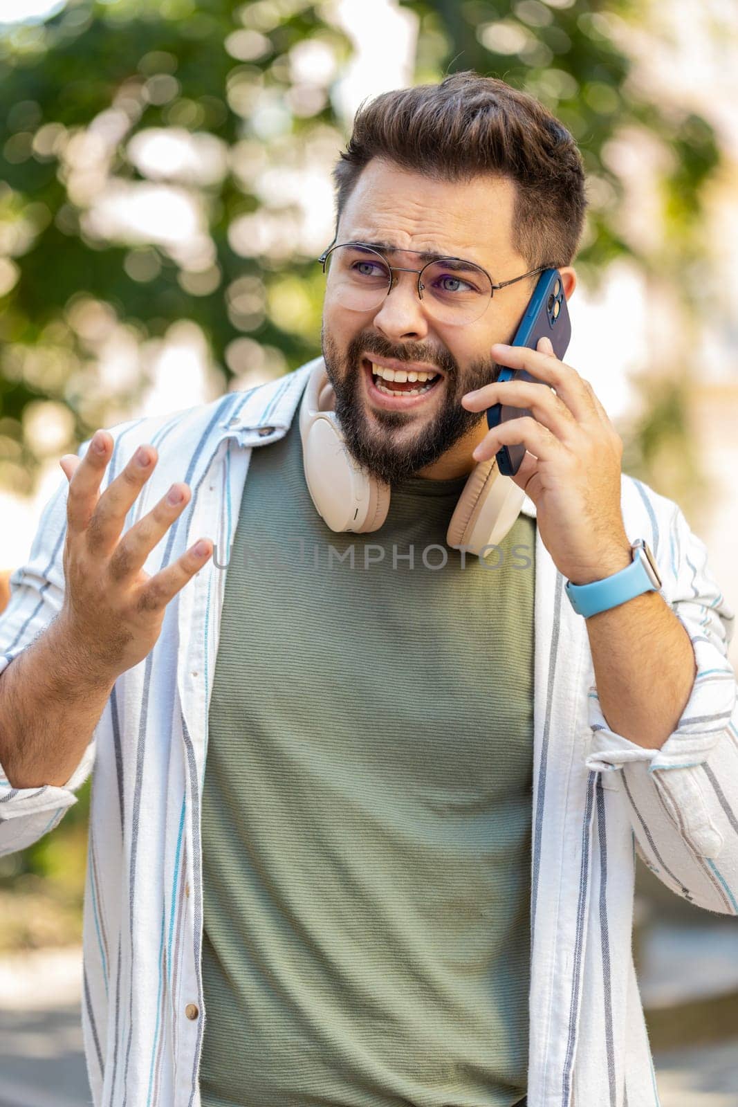 Angry adult Caucasian bearded man traveler arguing while talking on smartphone walking on the urban city street. Dissatisfied guy in glasses tourist shout into the phone, quarrels, expresses a problem