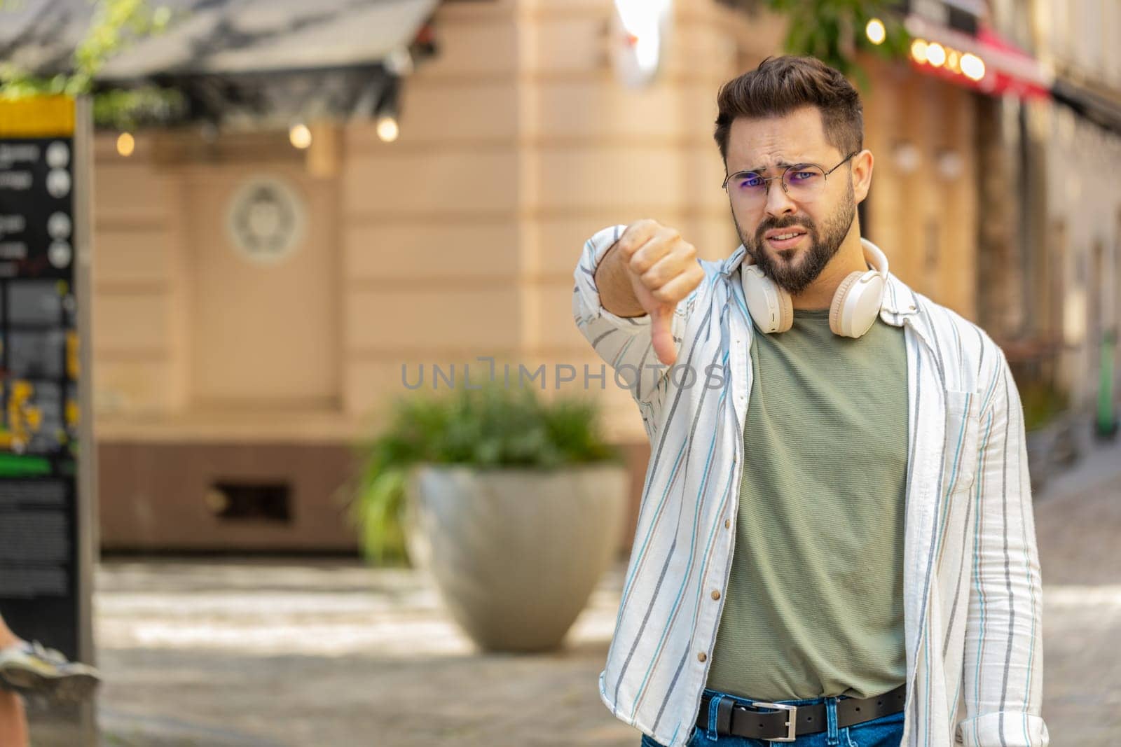 Dislike. Upset Caucasian young man showing thumbs down sign gesture, expressing discontent disapproval, dissatisfied bad work, mistake outdoors. Displeased guy tourist standing on urban city street.