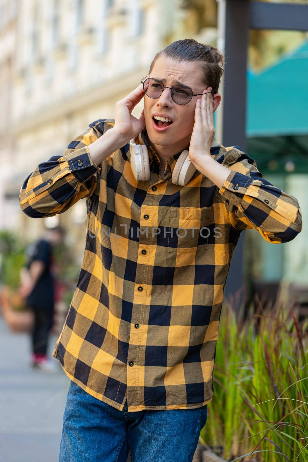 Displeased exhausted Caucasian man male rubbing temples to cure headache problem suffering from tension migraine stress. Young guy tourist in sunglasses grimacing, pain standing on city urban street.