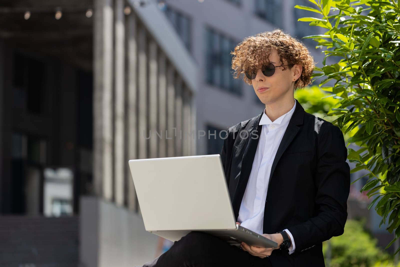 Caucasian young businessman investor employer entrepreneur sitting outside office building opening laptop and working on business project in city. Freelancer guy man in elegant suit and sunglasses.
