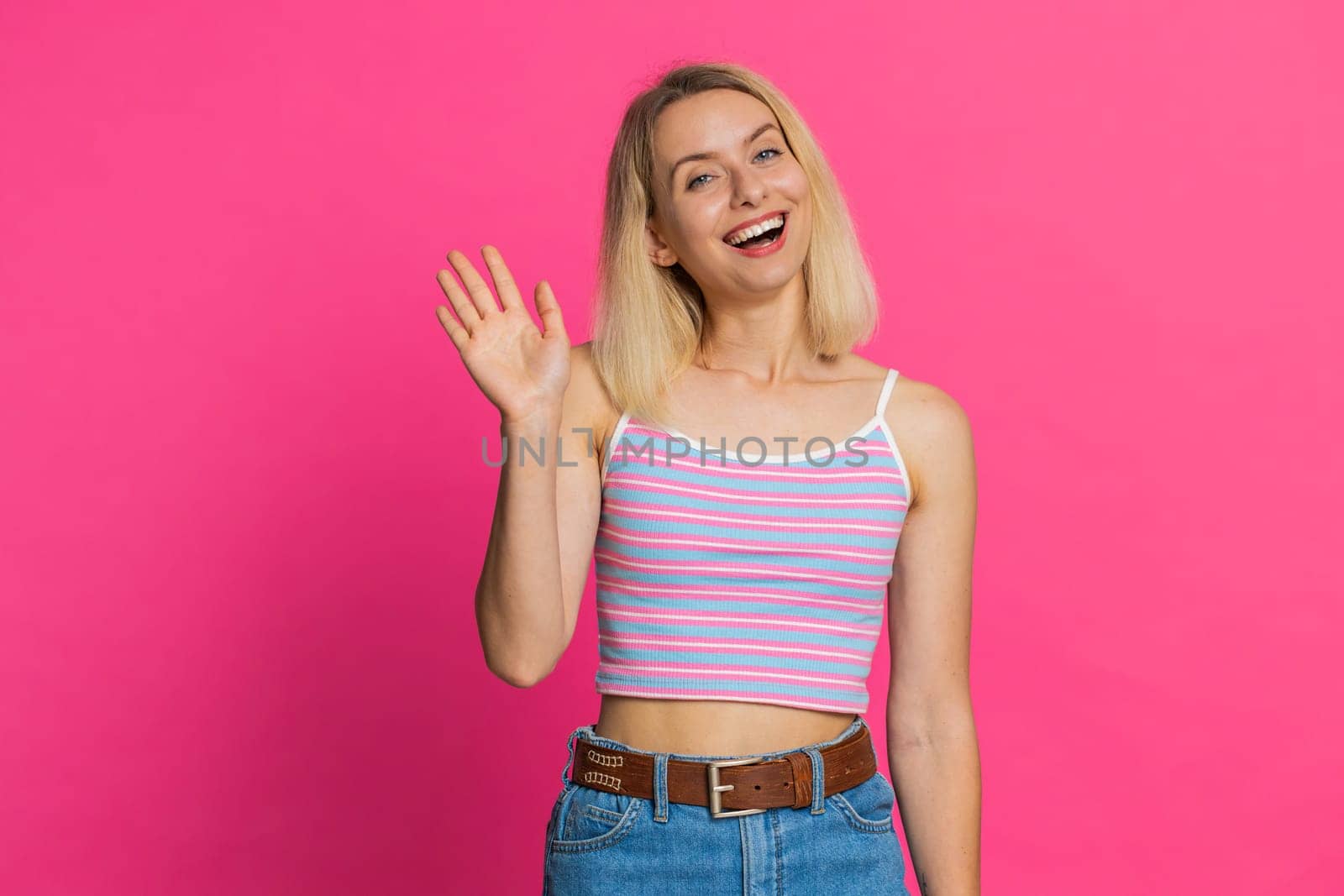 Hello. Young Caucasian woman smiling friendly at camera, waving hands gesturing hi greeting or goodbye welcoming with invitation hospitable expression. Pretty girl isolated on pink studio background