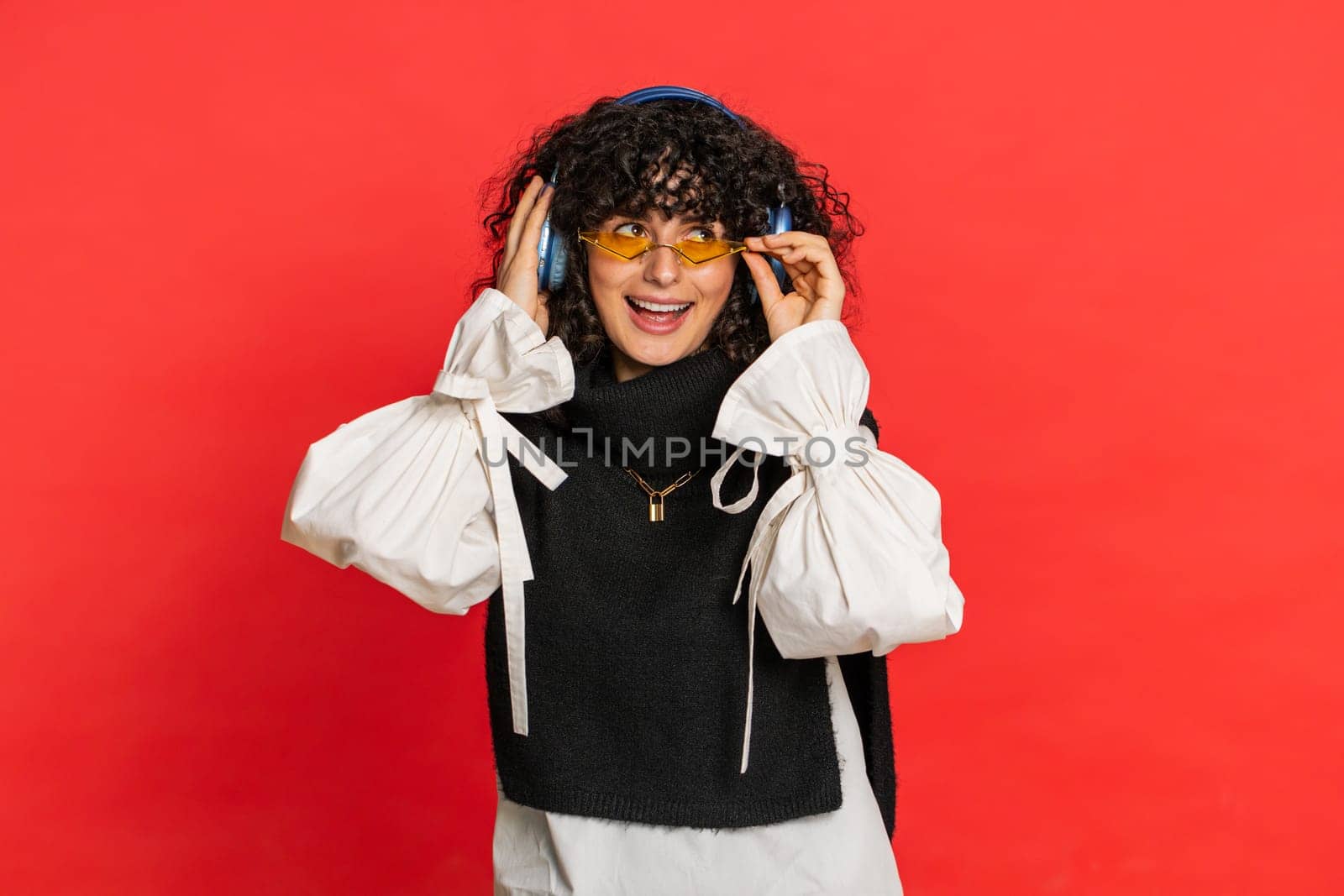 Caucasian young woman with curly hair listening music on headphones, dancing disco fooling around having fun expressive gesticulating hands. Music app advertisement. Girl isolated on red background