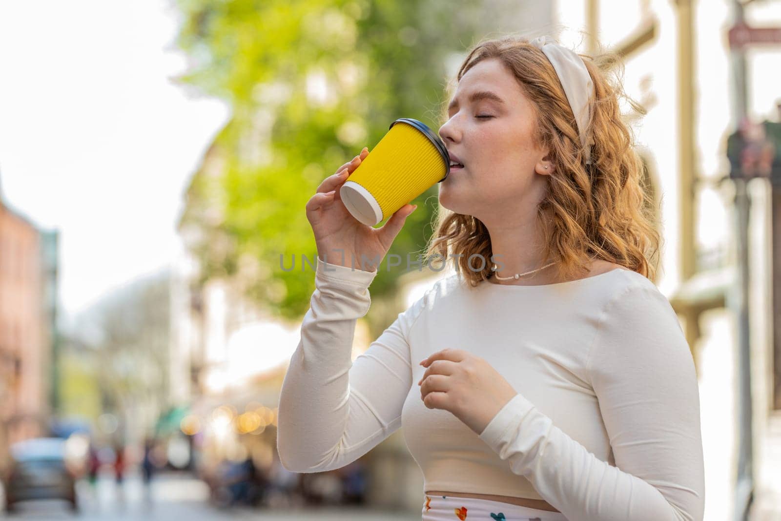 Happy redhead young woman enjoying morning coffee hot drink and smiling outdoors. Relaxing, taking a break. Girl drinking coffee to go standing in urban city center street. Town lifestyles outside.