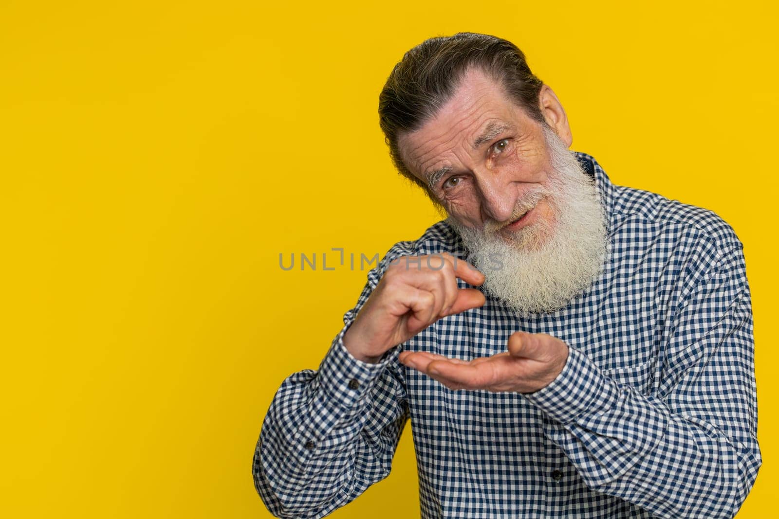 Need some more, please give me. Senior old man showing a little bit gesture with sceptic smile showing minimum sign measuring small size begging help. Elderly grandfather on yellow background indoors