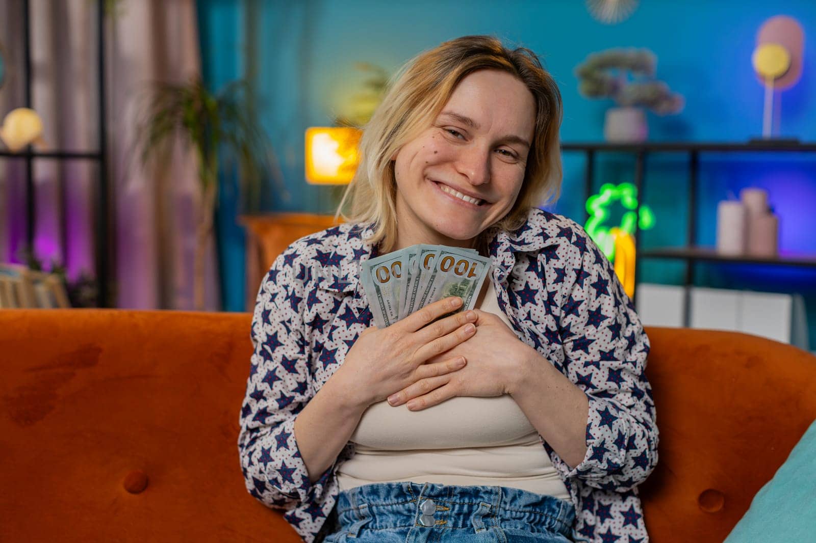 Successful rich Caucasian woman holding waving dollar bill money fan sitting on couch at home room. Happy excited girl winning online casino lottery game planning vacation calculating budget at home.