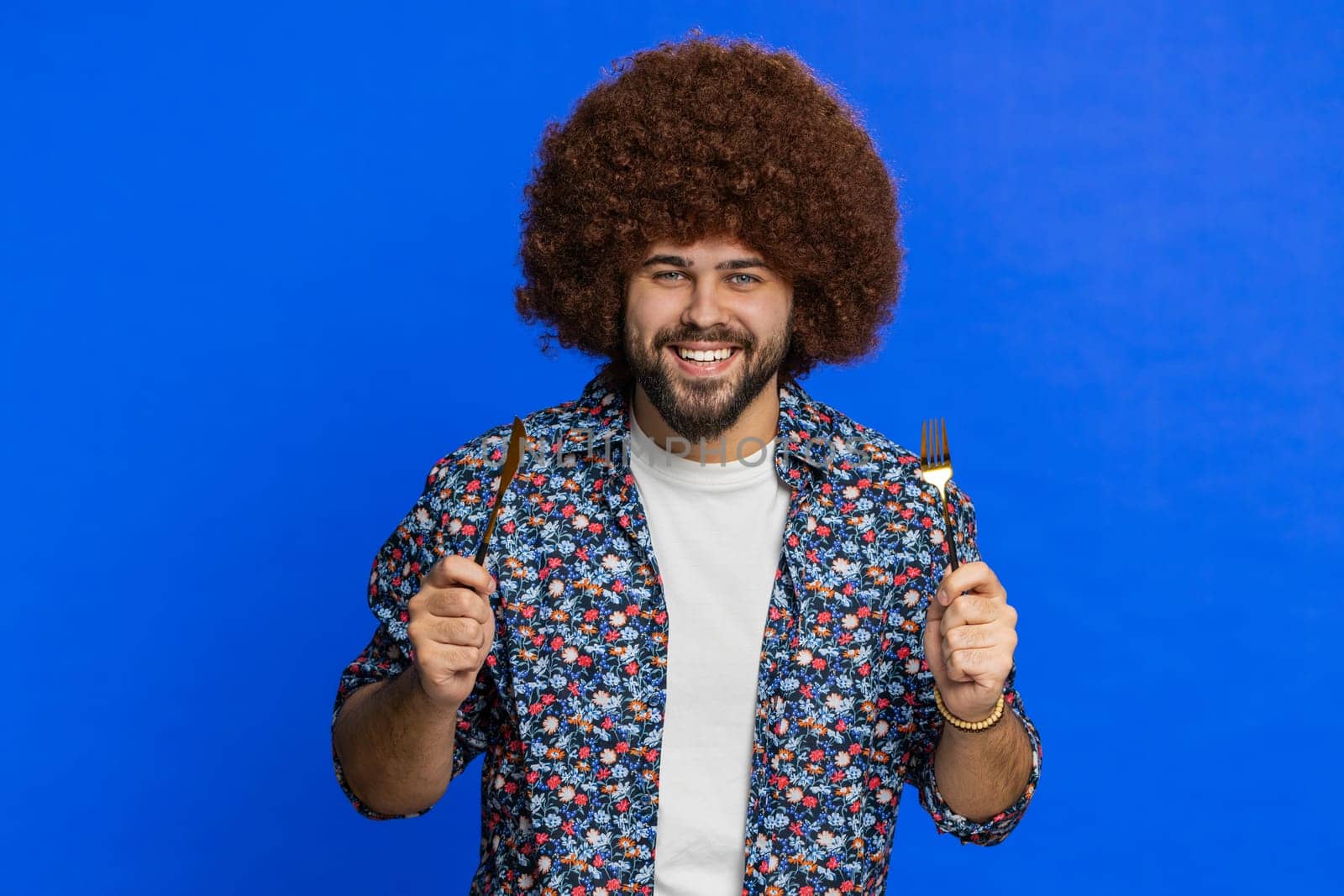 Ready to eat. Hungry man with Afro hairstyle wig waiting for serving dinner dishes with with restlessness holding cutlery fork knife, will appreciate delicious restaurant meal. Guy on blue background