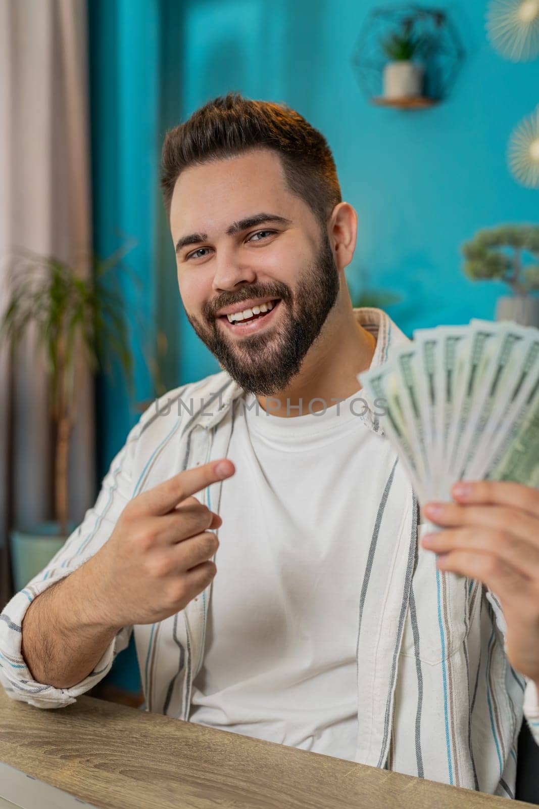 Young businessman guy holding fan of cash money dollar banknotes showing pointing on banknotes, celebrate, success career, online income, wealth at home office desk. Remote distant working. Vertical