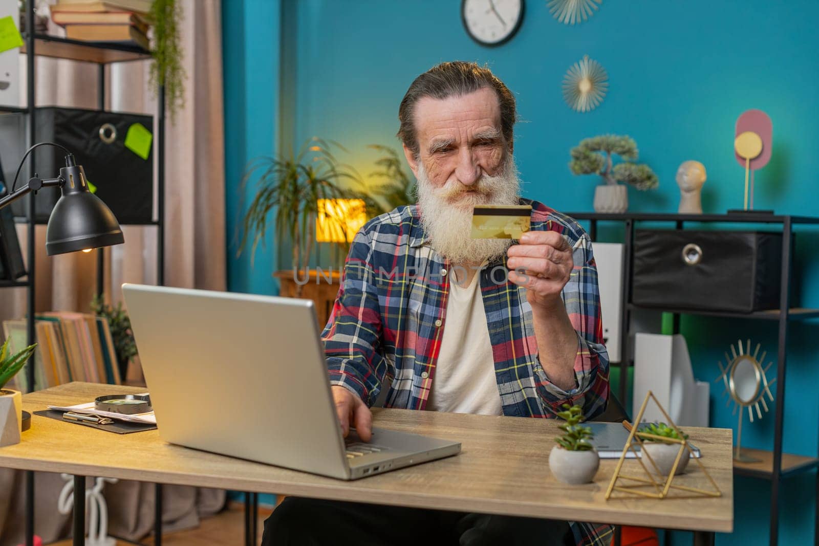 Happy Caucasian senior man using bank credit card and laptop for online shopping payments at home office. Business transaction. Smiling grandpa freelancer purchases with netbook E-banking app service.