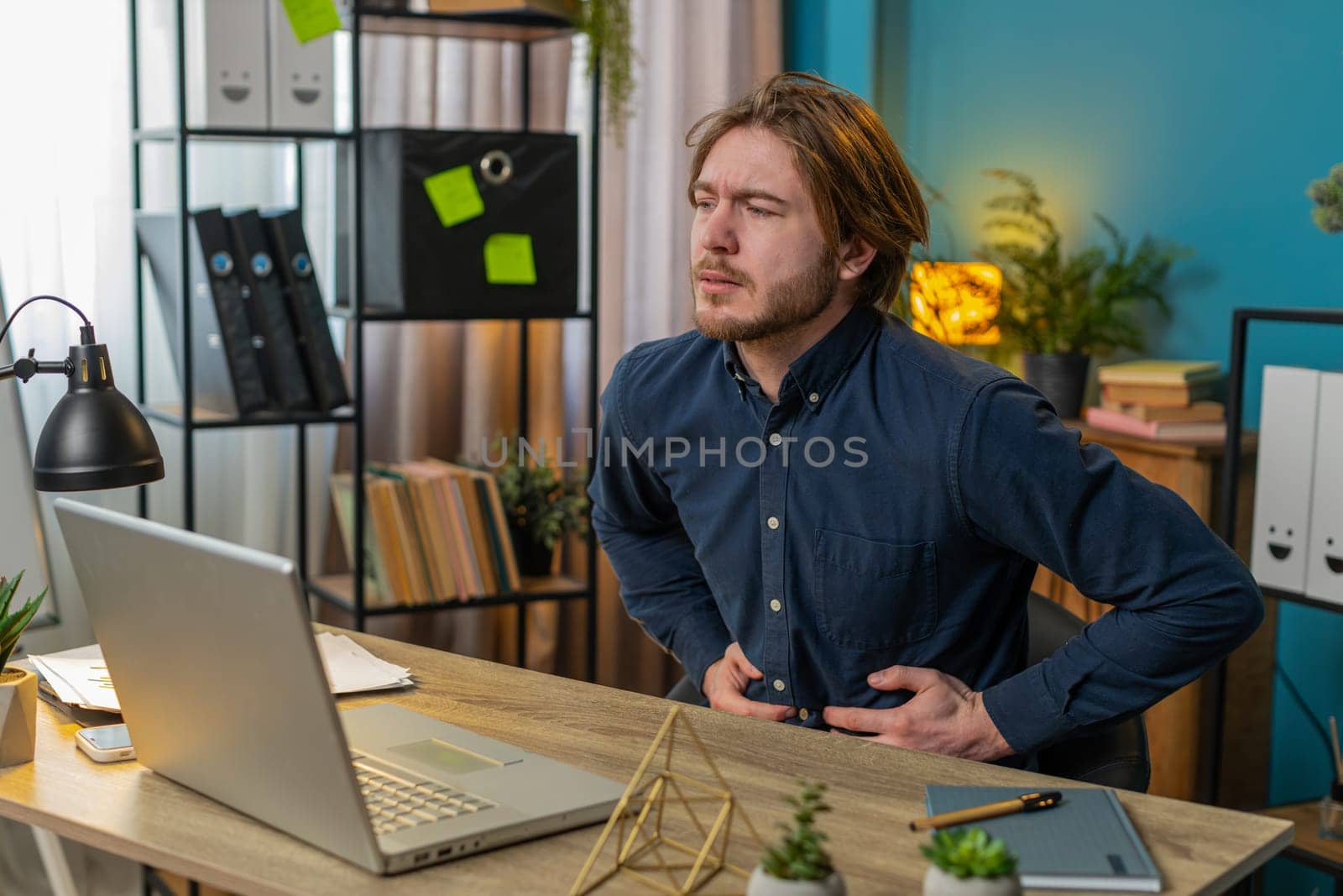 Disappointed Caucasian freelancer feeling overeating during work process on laptop in home office. Upset businessman freelancer guy man touching stomach and experiencing discomfort and sharp pain.