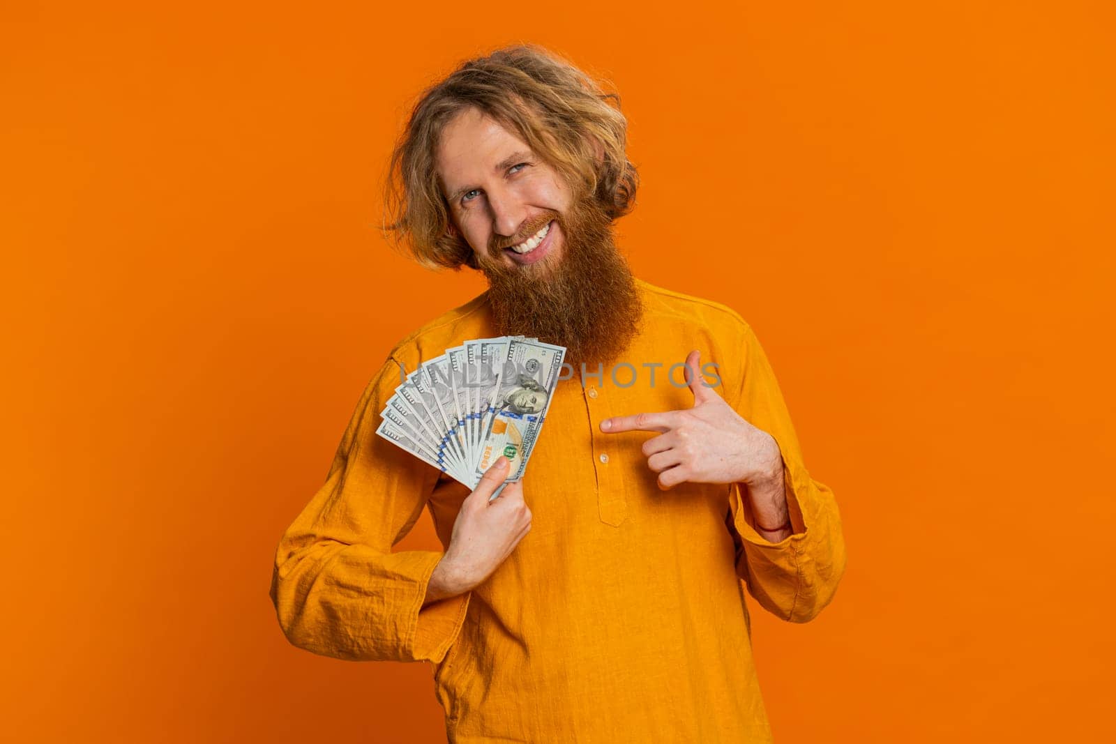 Rich pleased happy Caucasian man waving money dollar cash banknotes bills like a fan, success business career, lottery winner, big income wealth. Young adult guy isolated on orange background indoors