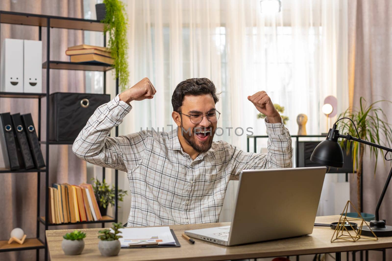 Happy amazed Indian businessman working on laptop shocked by sudden victory, game winning lottery goal achievement, good career news. Excited Hispanic guy freelancer clenching fists at home office.