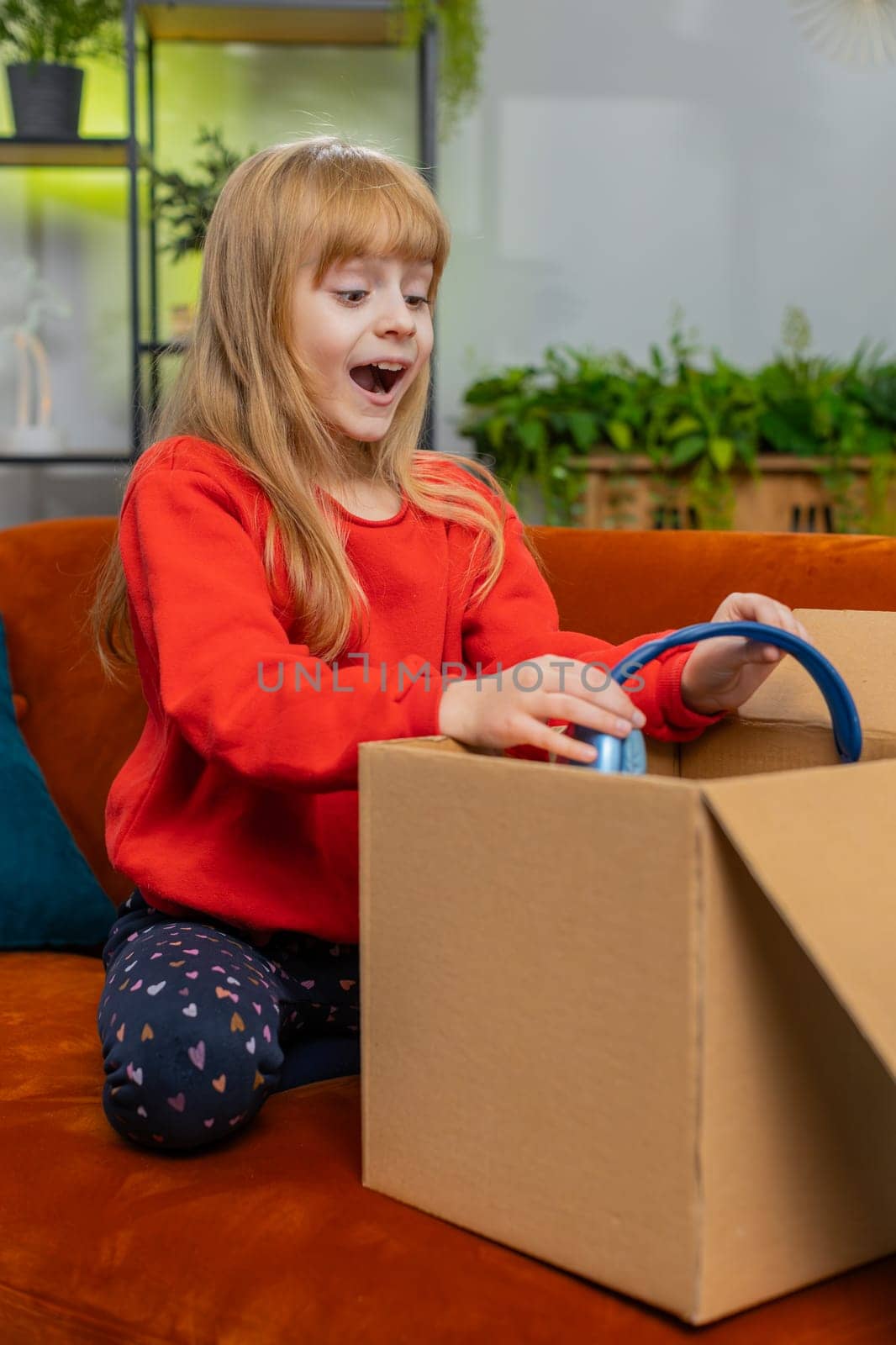 Happy young child girl unpacking delivery parcel. Smiling satisfied teenager kid shopper online shop customer opening cardboard box receiving purchase headphones gift by fast postal shipping. Vertical