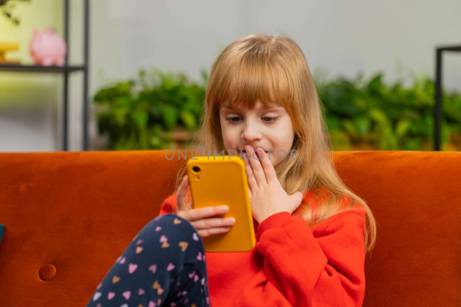 Young child girl texting share messages content on smartphone social media applications online watching relax movie. Female teenager little kid uses mobile phone at home in living room sitting on sofa