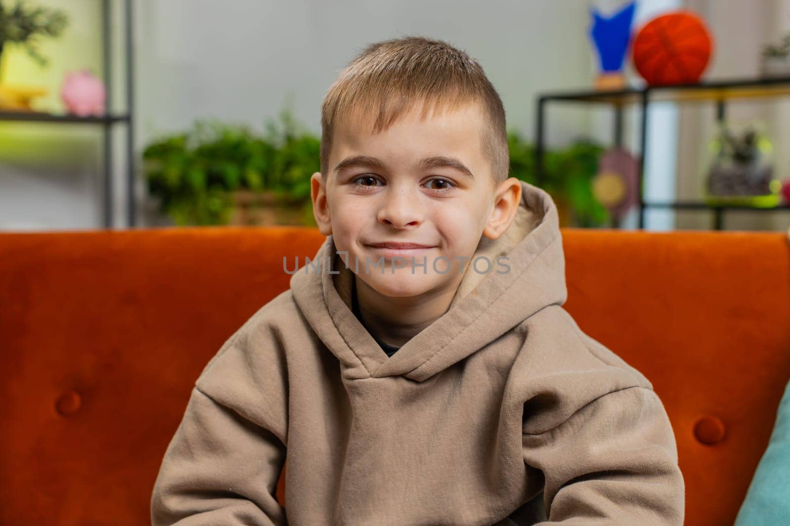Close-up portrait of happy smiling cheerful Caucasian preteen school boy 8 years old. Young lovely blonde little child kid looking at camera at home play living room apartment sitting on orange sofa