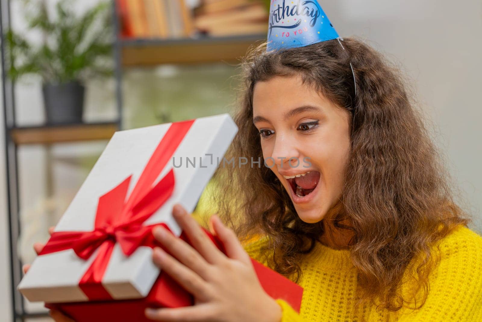 Happy 15 years old child girl wears festive birthday cap hat hold gift box with ribbon congratulating. Teenager female kid celebrating party event opening delivery greeting present at home room