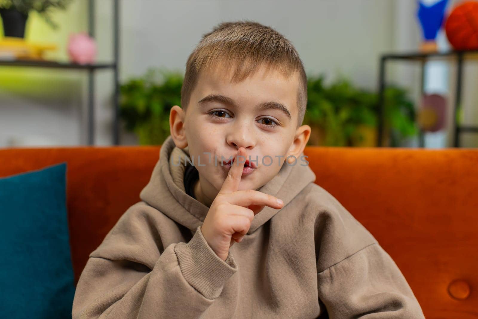 Shh be quiet please. Preteen little child boy presses index finger to lips makes silence hush gesture sign do not tell gossip secret. Teenager male little kid at home apartment play room sits on couch