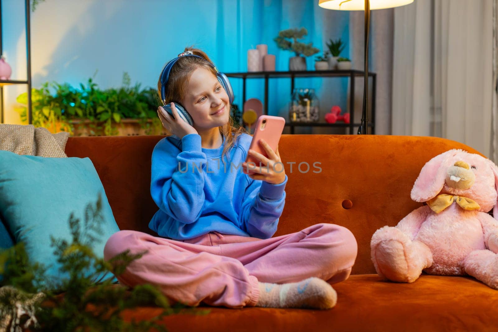 Happy overjoyed teenager girl kid in wireless headphones relaxing sits on couch at evening home room choosing listening favorite energetic disco dancing music. Children weekend leisure activities