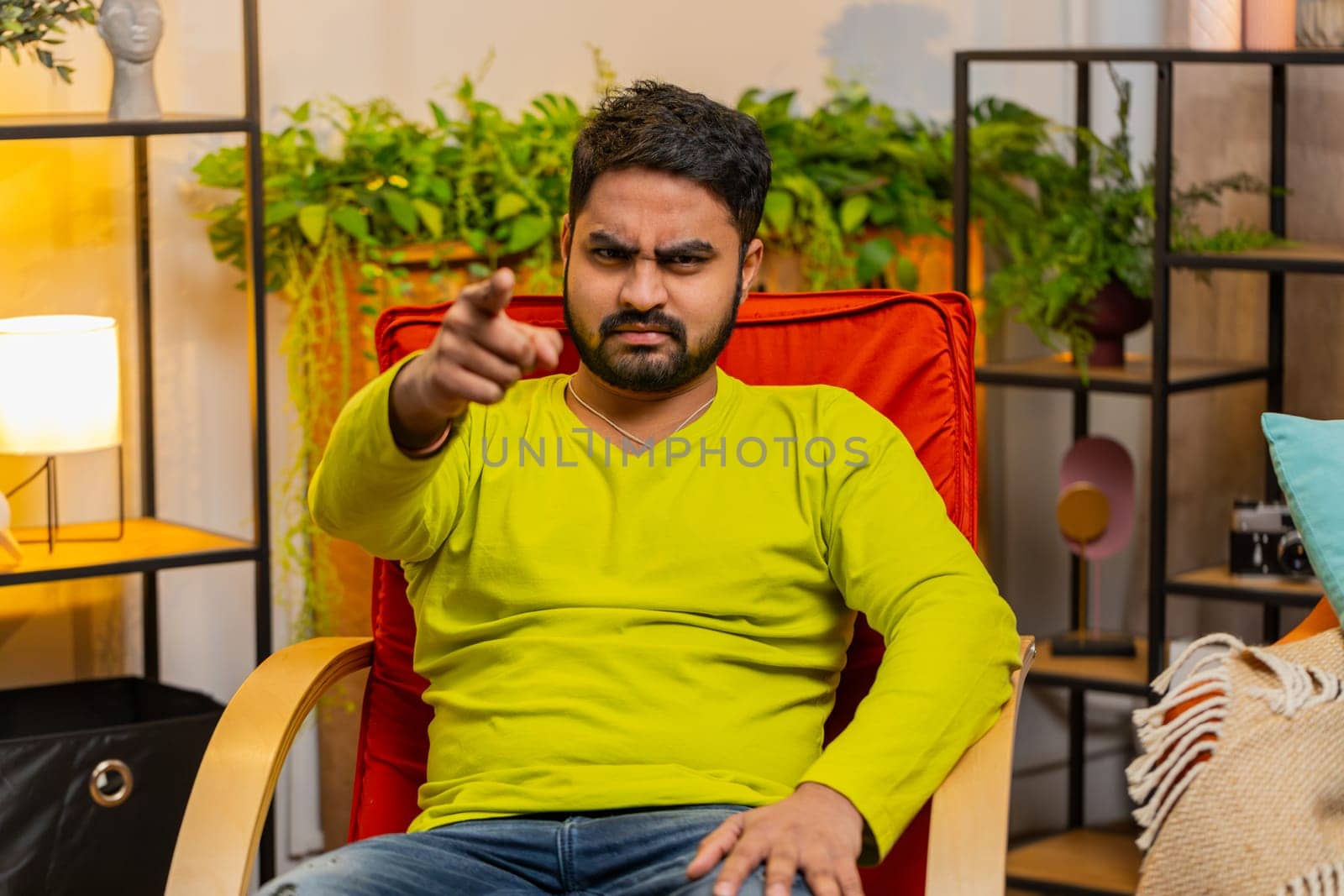 Hey you. Strict unhappy Indian man pointing hand finger to camera, looking angry, making choice, choosing guilty, noticing suspicious, accusing for mistake at home room apartment. Guy sits on chair