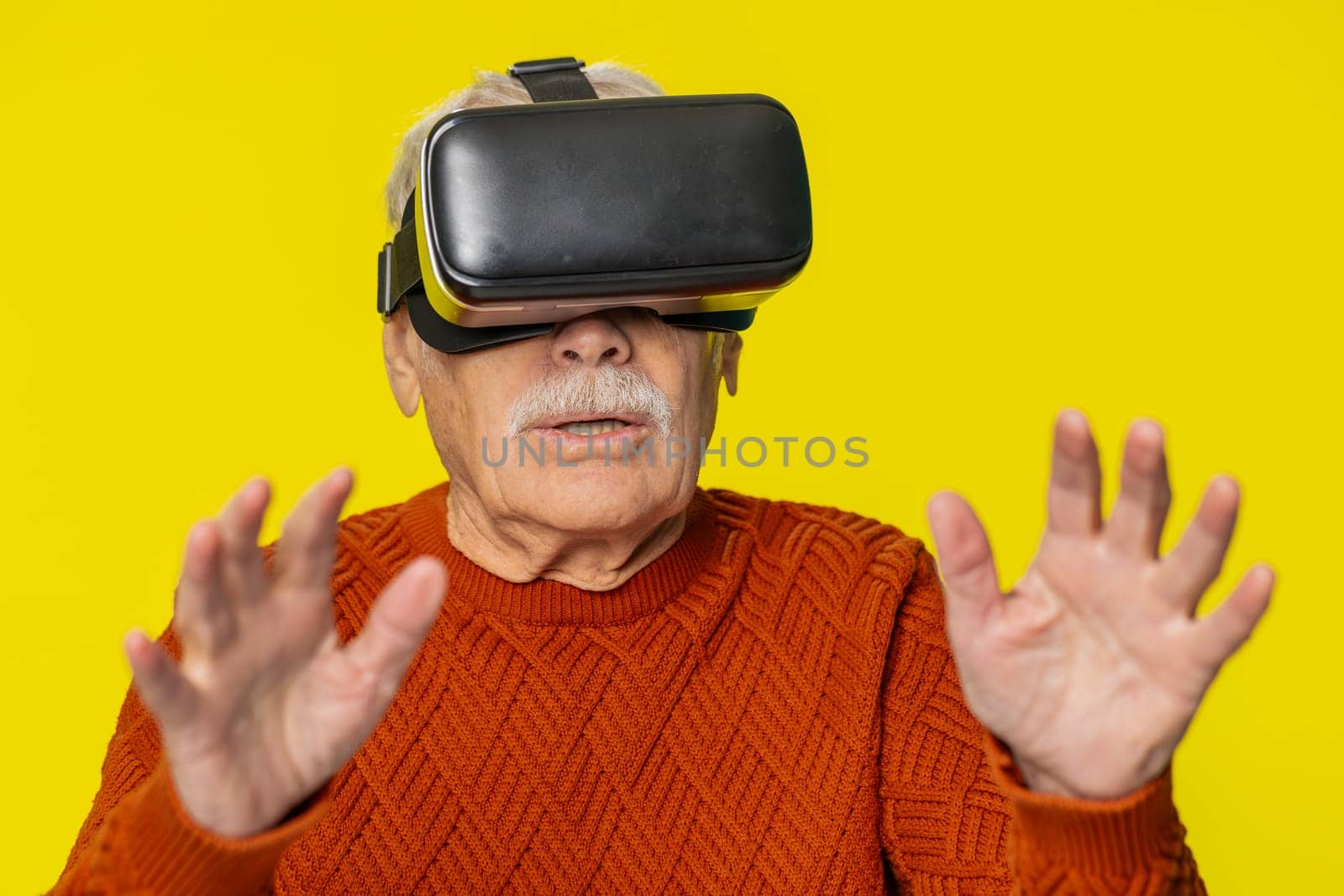 Excited happy senior old man using headset helmet app to play simulation game. Watching virtual reality 3D 360 video. Elderly grandfather in VR goggles isolated on yellow background. Future technology
