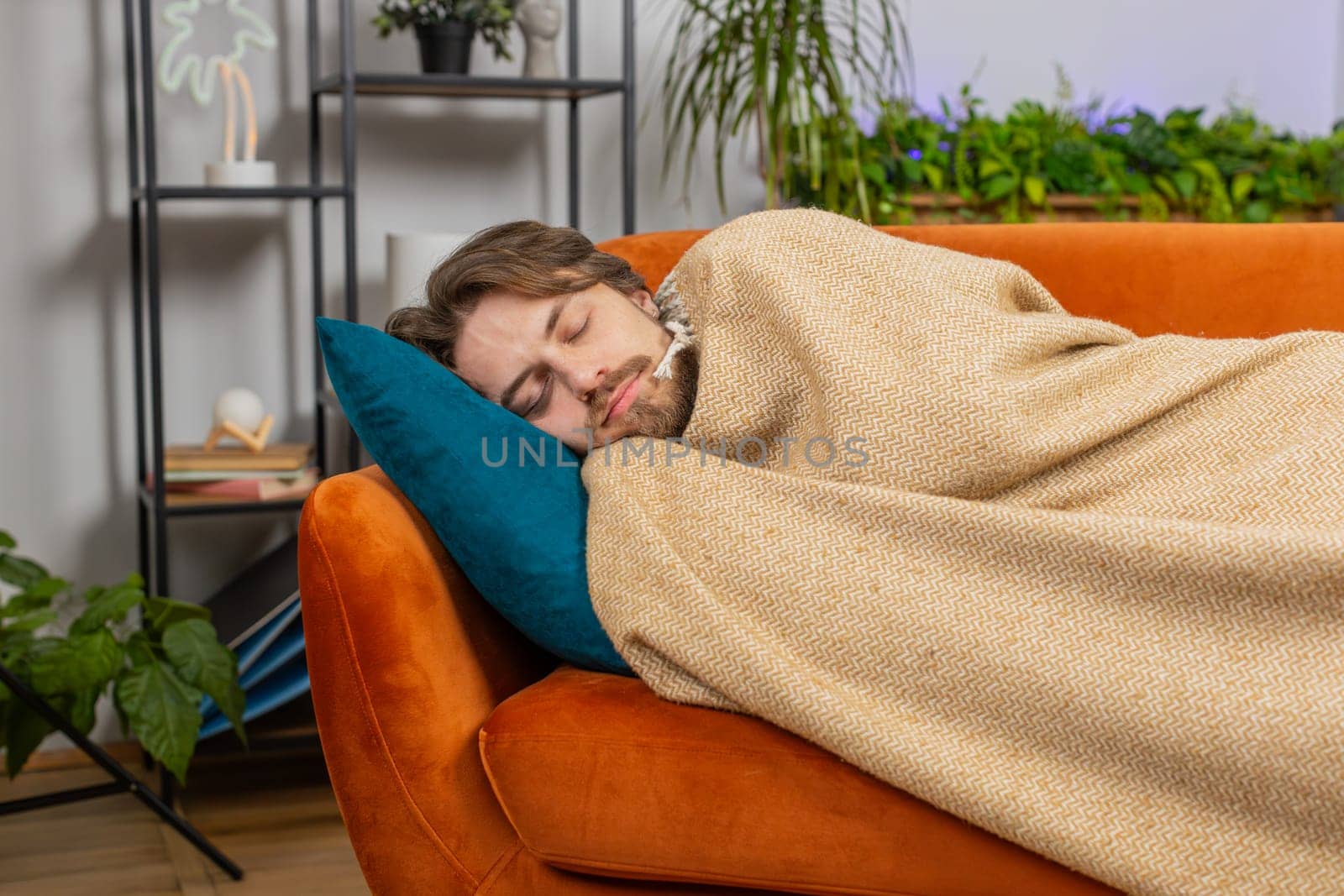 Tired young man lying down in bed taking a rest at home apartment room. Carefree Caucasian guy napping, falling asleep on comfortable orange couch with pillows. Closed his eyes enjoy daytime nap alone