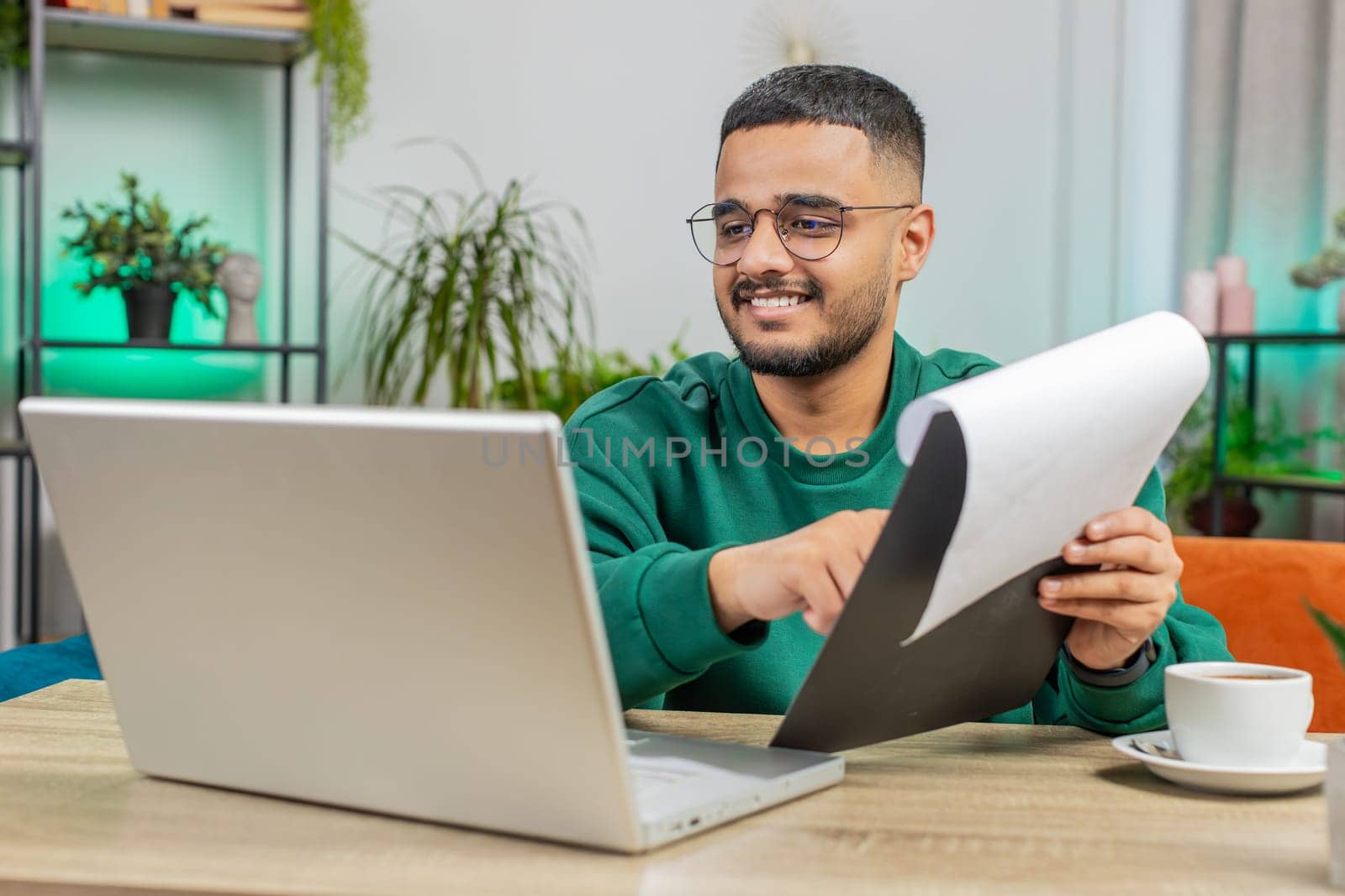 Happy Indian man freelancer work with documents from home analyzing information read business papers prepare financial report. Smiling guy winner shocked by sudden good victory success news on laptop