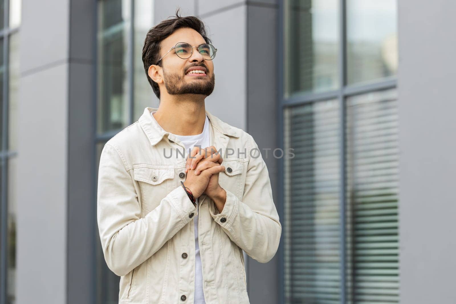 Young Indian man praying with closed eyes to God asking for blessing, help, forgiveness outdoors. Arabian confident guy clasping hands wishing luck in downtown city street. Business people lifestyles