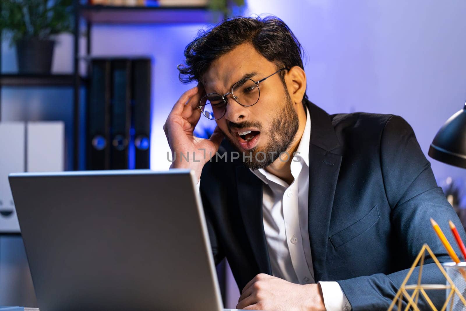 Tired ill Indian businessman suffering from headache problem tension and migraine, stress at home office desk workplace. Bearded freelancer broker worker man works on laptop notebook. Overworking