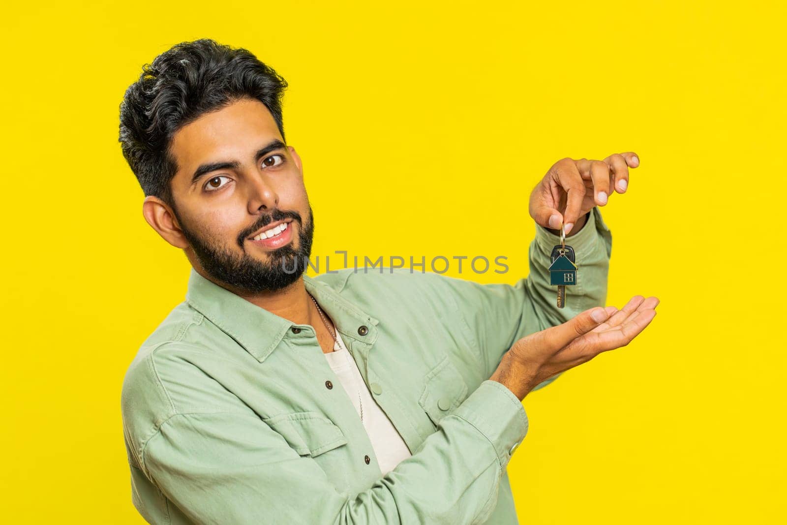 Indian young man real estate agent lifting hand showing the keys of new home house apartment, buying or renting property, mortgage loan, insurance. Arabian guy isolated on yellow studio background