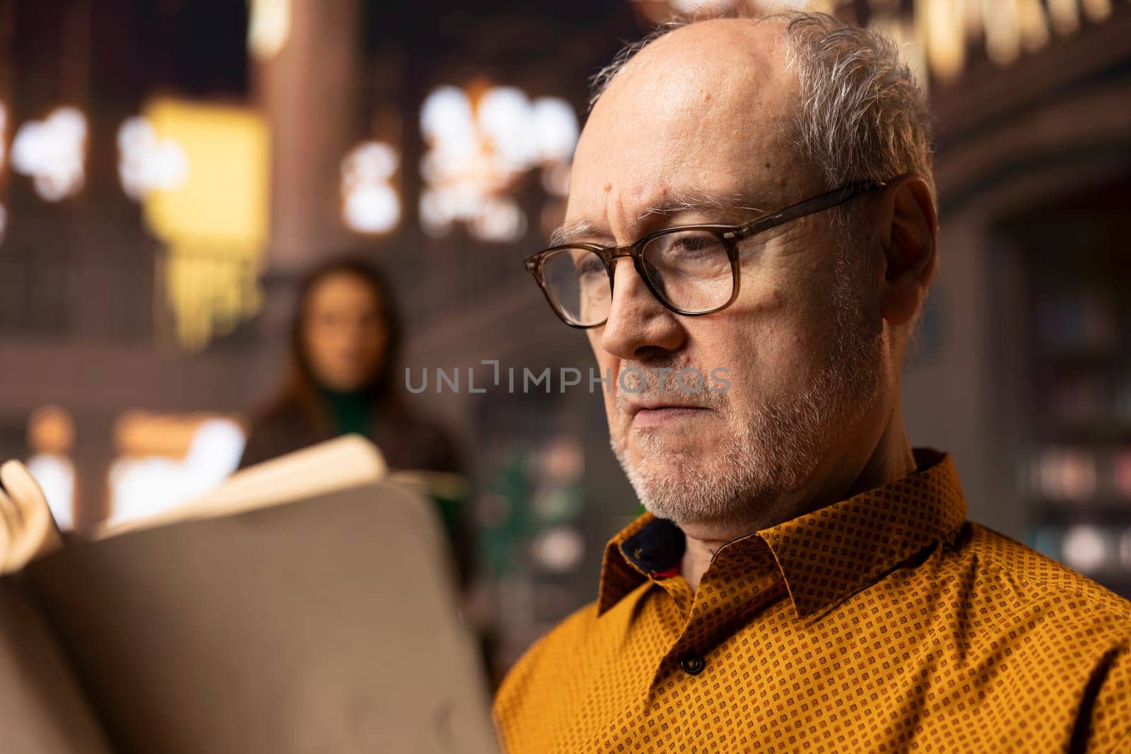Retired man enjoys book reading at a public library and for leisure activity, feeling relaxed and increasing his knowledge during retirement. Happy calm person with eyeglasses, recreation.