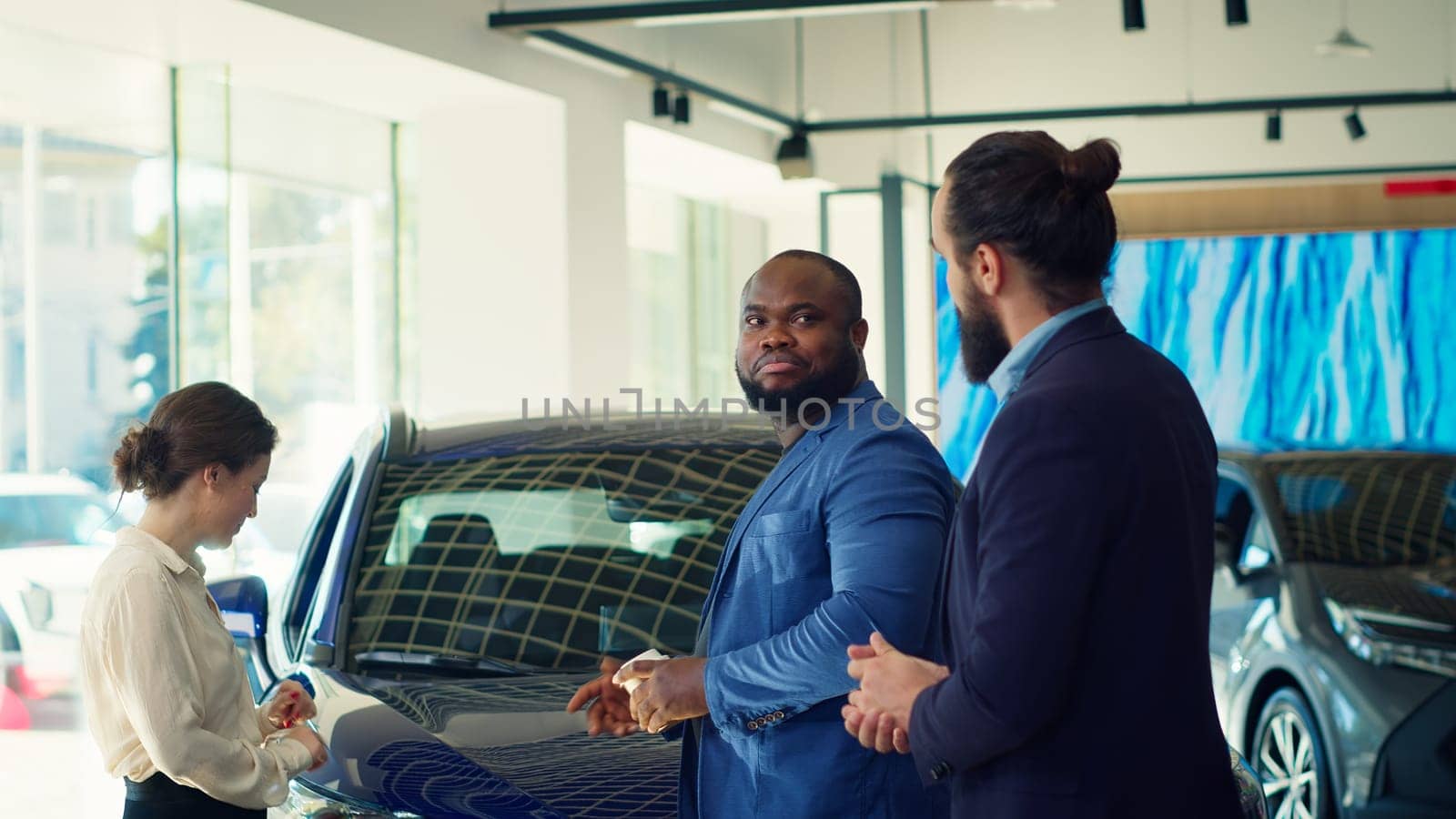 Clients arriving in car dealership, being greeted by friendly car salesman ready to showcase available vehicles to clients. Customers walking in showroom, being welcomed by agent, camera A