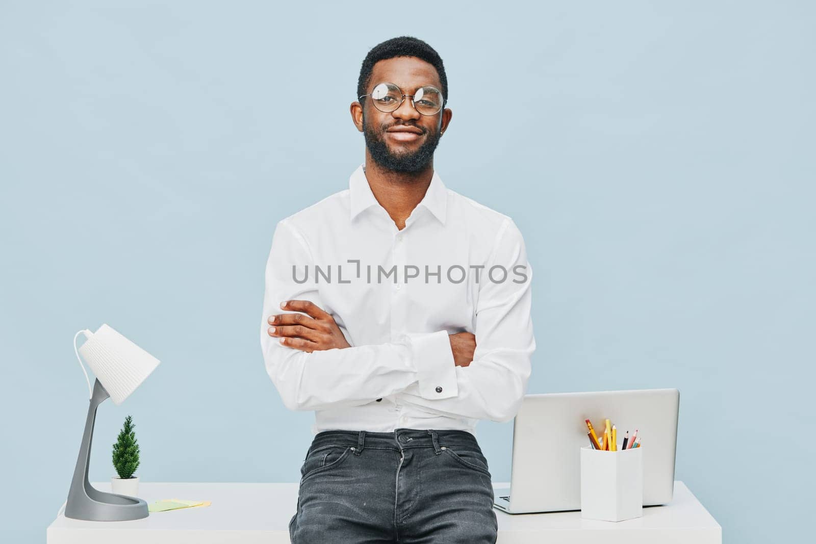 Friendly young man with glasses standing at sleek office desk with crossed arms, smiling, contemporary workspace with laptop, white lamp, and stationery, professional atmosphere, light blue backdrop, positive mood