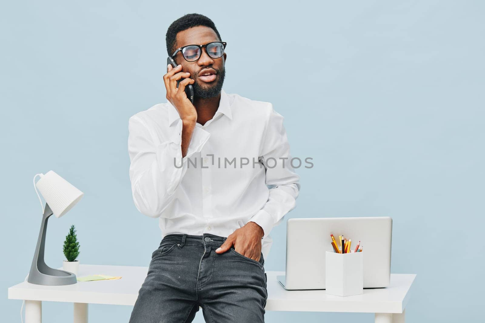 Confident young man in formal clothing engaging in a phone call inside a contemporary office environment, standing near a desk with a laptop and stationery, conveyance of business communication, professional atmosphere, workspace, office lifestyle