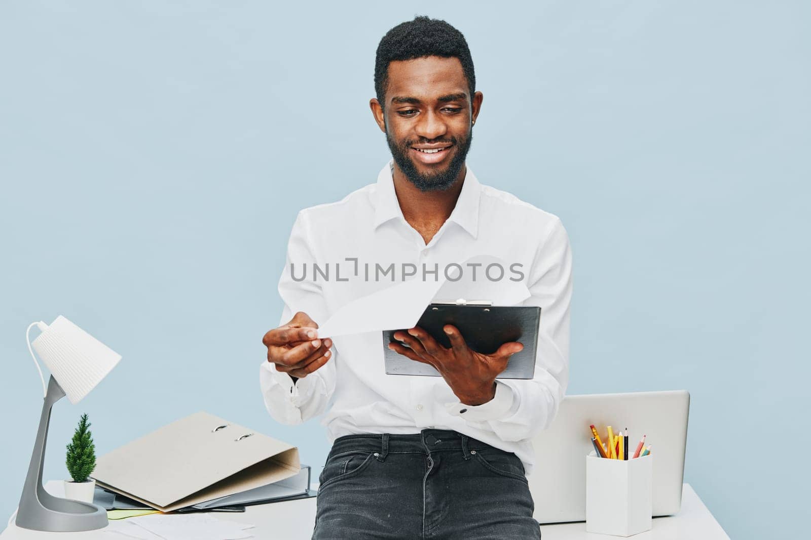 Happy man dressed in formal white shirt using tablet in a professional office setting, surrounded by office supplies and a laptop, positive mood, modern workspace, indoor shoot, business lifestyle concept.
