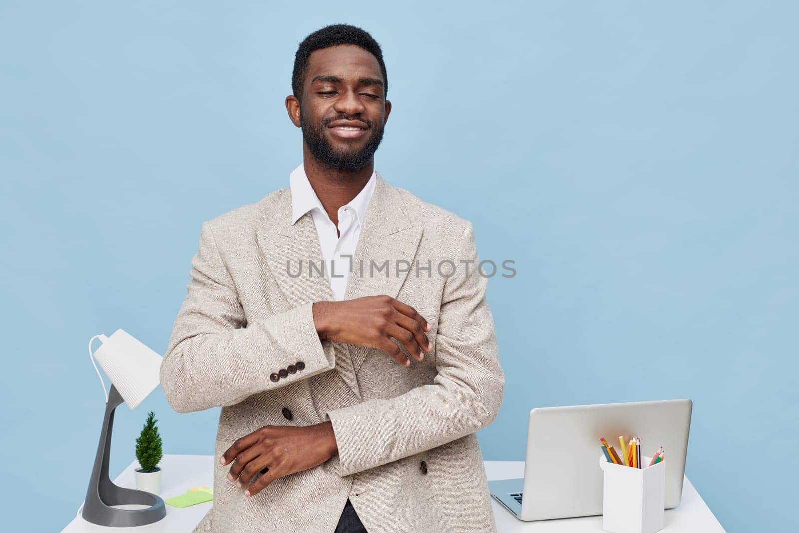 Portrait of a confident young African man in a stylish beige blazer and white shirt, smiling and looking at the camera in a modern office environment with a laptop, stationery, and a minimal pine plant, emphasizing professionalism and success. Concept: professional confidence