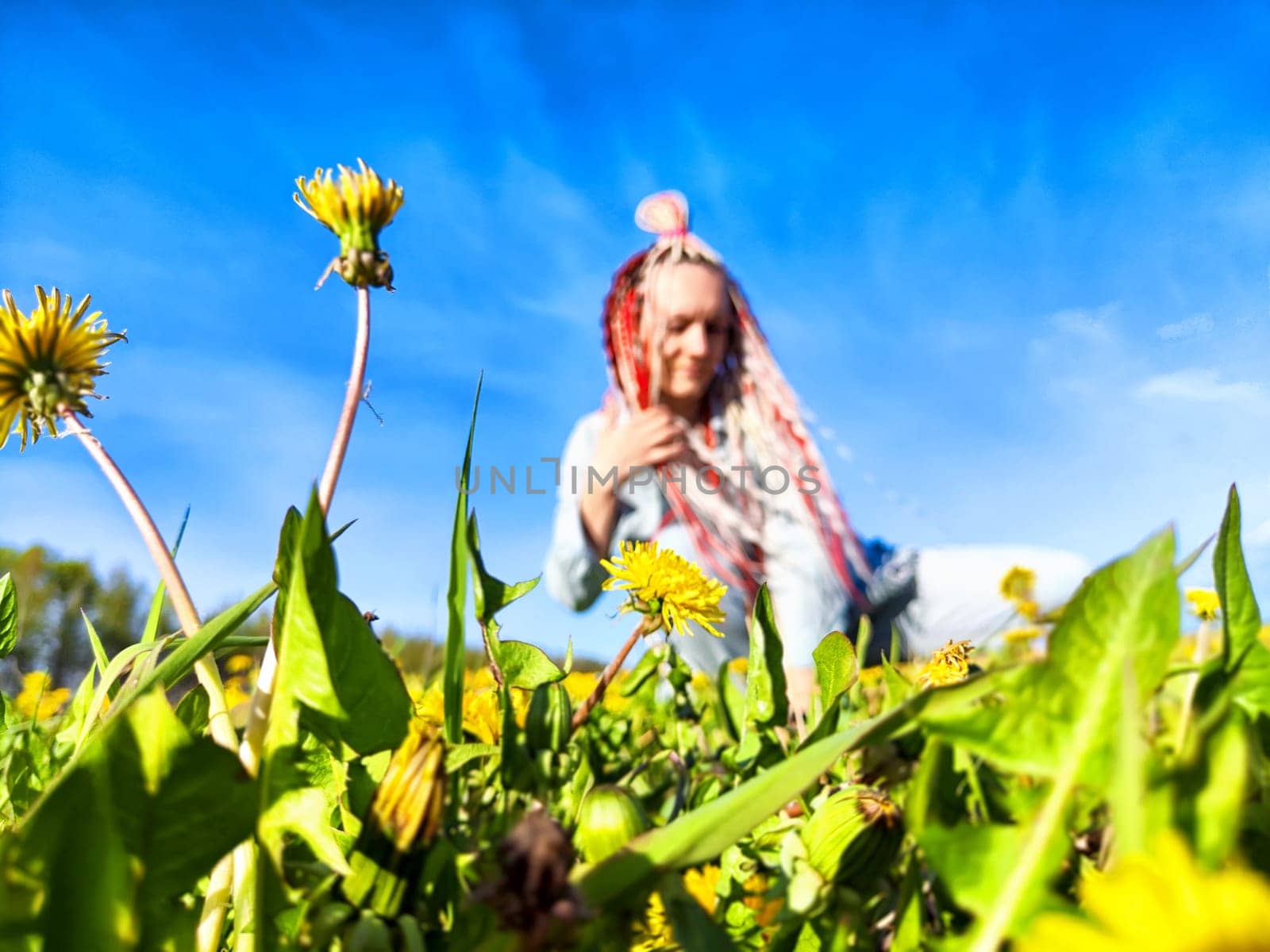 A person with bright red and white braids interacts playfully with dandelions in a sunny field against a blue sky.
