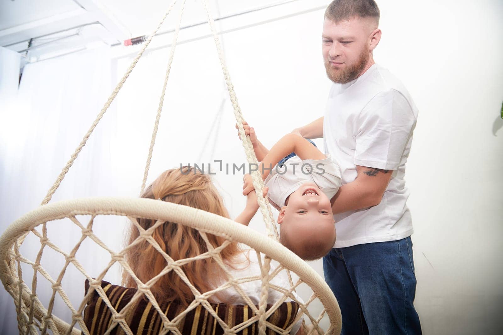 Father lifts his smiling child while the mother watches happily from a swinging chair, creating joyful family memories in a bright room.
