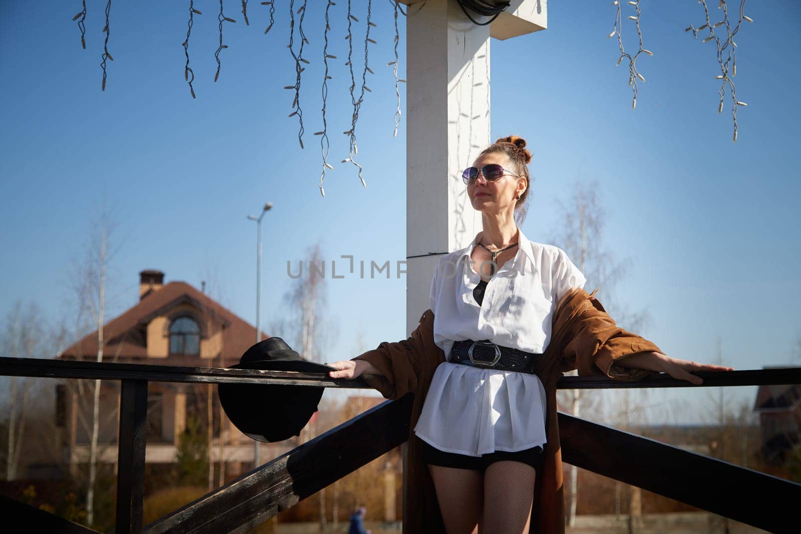 A woman stands with confidence on a wooden deck, dressed in a fashionable outfit, enjoying a sunny day in a tranquil suburban setting.