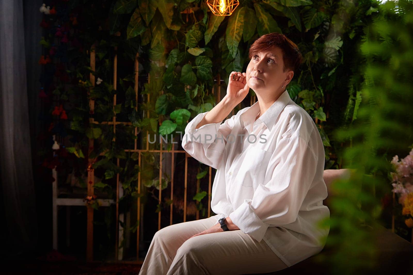A middle-aged woman sits thoughtfully in a cozy room surrounded by lush plants, showcasing her relaxed lifestyle.