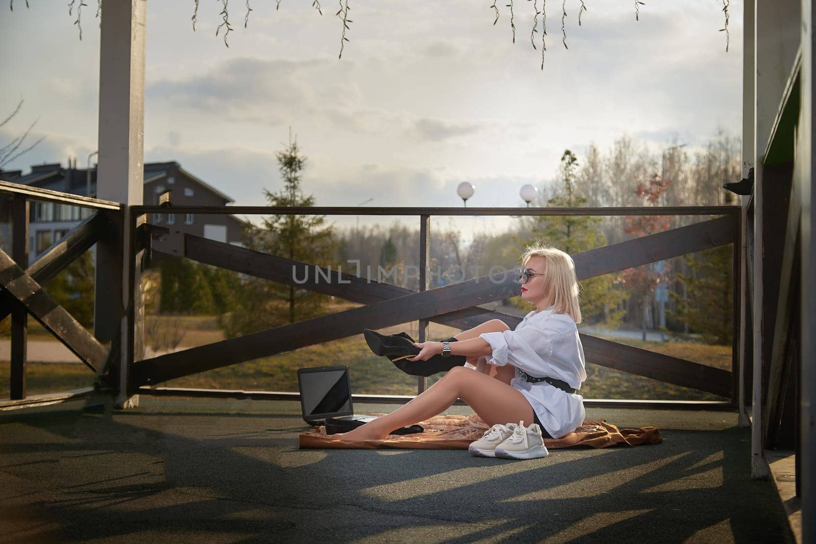 A young woman adjusts her shoes while seated on a cozy balcony, preparing for outdoor work on a bright, sunny day.