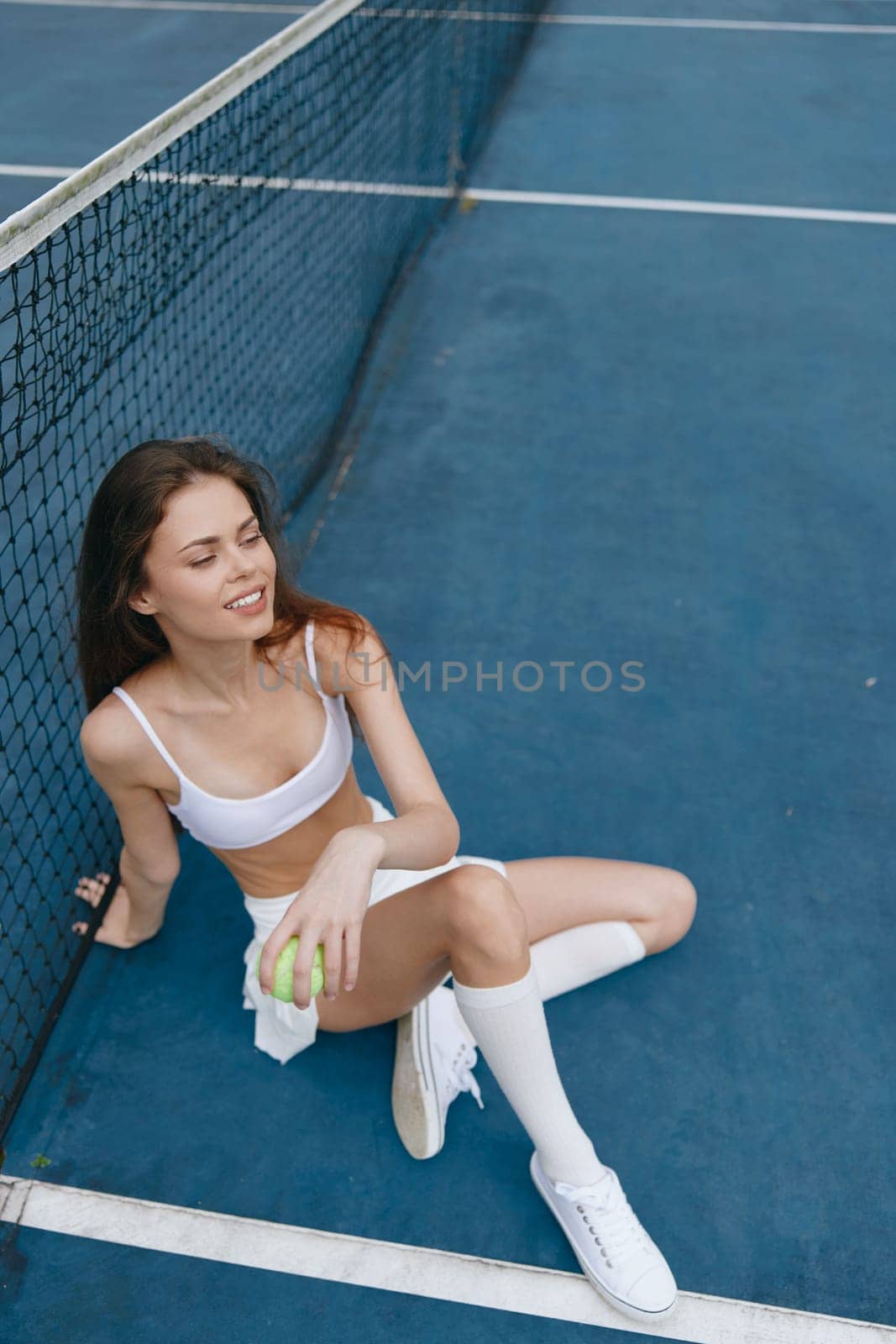 Athletic young woman in white sportswear, smiling while sitting on a tennis court with a tennis ball, embodies a joyful and energetic sports lifestyle against a vibrant blue background