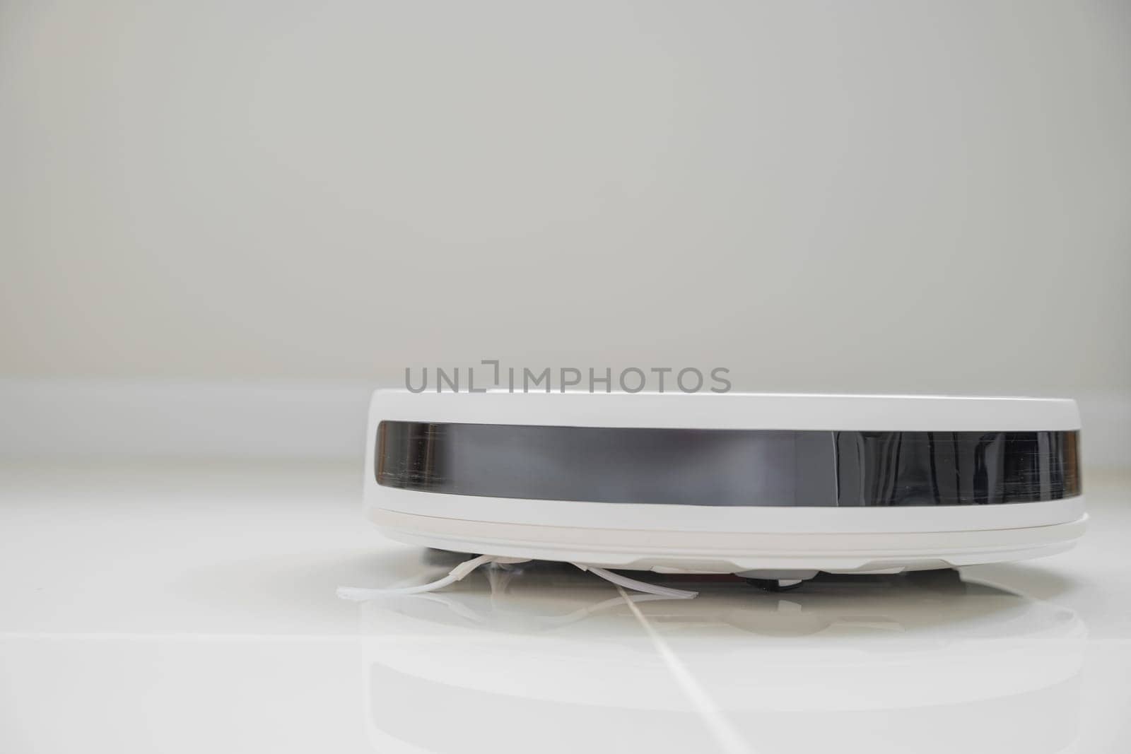 White automatic floor cleaner with side brush on tile flooring, featuring front black sensor strip. Robot vacuum cleaner efficiently cleaning floor, illustrating smart home technology for dust removal