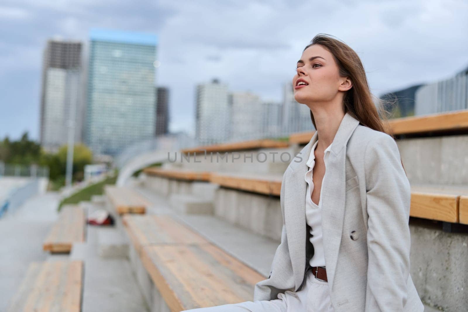 Thoughtful woman in light gray business suit sitting on outdoor bleachers with modern city skyline blurred, expressing calm confidence and autumn atmosphere, business concept